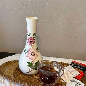 May include: A white ceramic vase with pink floral designs and green leaves sits on a woven tray. A glass teacup filled with tea and tea bags are also on the tray. The tea bags are labeled "Bigelow Constant Comment".