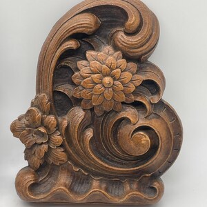 May include: A carved wooden decorative element featuring floral and scroll designs. The piece is stained in shades of brown, with detailed carvings of flowers and swirling patterns. The overall shape is asymmetrical, with a curved top and a flat base.