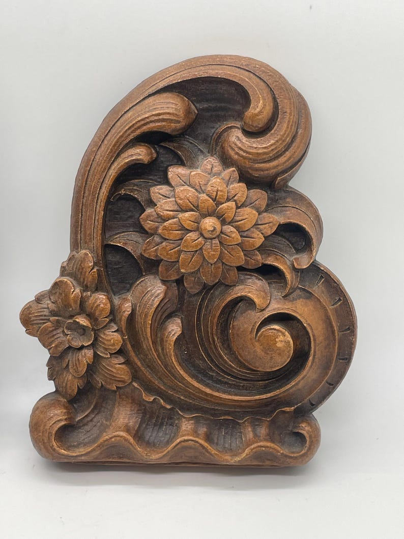 May include: A carved wooden decorative element featuring floral and scroll designs. The piece is stained in shades of brown, with detailed carvings of flowers and swirling patterns. The overall shape is asymmetrical, with a curved top and a flat base.