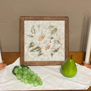 May include: A decorative arrangement featuring a framed tile with a floral design, two white candles, a bunch of green grapes, and a green pear. The tile has a wooden frame and a white background with a daisy-like flower design.