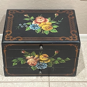 May include: A black wooden jewelry box featuring hand-painted floral arrangements in shades of pink, yellow, and blue on the top lid and the front drawer. The box includes two drawers with small silver-toned knobs and decorative brown border trim.