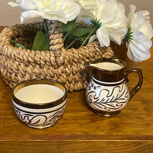 May include: A small, white ceramic creamer and matching bowl with gold trim and a black floral design. A woven basket with white flowers is in the background. The creamer has a handle and spout.