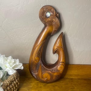 May include: A brown wooden fish hook carving featuring intricate spiral patterns and a single iridescent abalone shell inlay at the top. The hook has a smooth, polished finish with deep grooves that highlight the detailed wood grain and carvings.