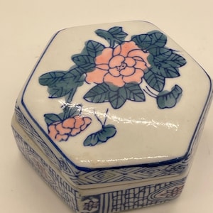 May include: A white, hexagonal ceramic trinket box with a lid. The lid features a pink floral design with green leaves. The box is trimmed with blue and has a patterned design around the sides.