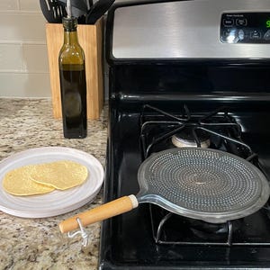 May include: A kitchen scene with a stainless steel tortilla warmer on a black stovetop. A plate with two tortillas sits next to it. An olive oil bottle and utensils are in the background.