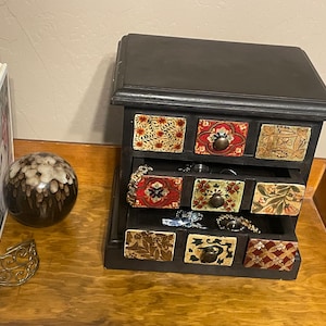 May include: A black wooden jewelry box with nine small drawers, each adorned with colorful, patterned panels. A gold-toned bracelet and a decorative sphere sit nearby. Books are visible in the background.