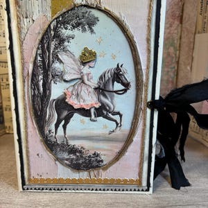 May include: A handmade journal with a fairy and horse illustration in an oval frame. The cover features a pink, gold, and cream color scheme with decorative trim and a black ribbon closure. The journal is a unique, artistic item.