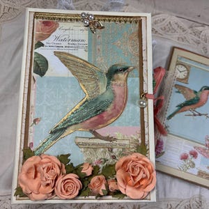 May include: A rectangular journal with a cream cover featuring a central illustration of a bird with green and pink wings. The cover is decorated with peach-colored paper roses, a gold-toned metal embellishment, and vintage-style text reading Waterman.
