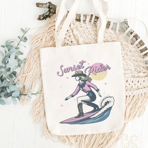 Beige tote bag displaying a Surfing Cowgirl graphic, showing a cowgirl on a surfboard with the text "Sunset Rider," ideal for beach outings and shopping
