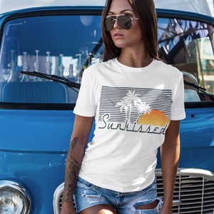 May include: A white t-shirt with a graphic of palm trees and a sunset. The text "Sunkissed" is printed below the graphic. The shirt is being worn by a person standing in front of a blue van.