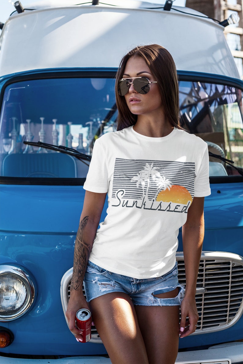 May include: A white t-shirt with a graphic of palm trees and a sunset. The text "Sunkissed" is printed below the graphic. The shirt is being worn by a person standing in front of a blue van.