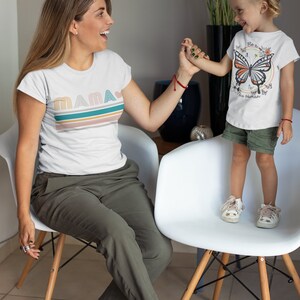 May include: A woman and a young girl are sitting on white chairs. The woman is wearing a white t-shirt with the word "MAMA" in colourful letters and green trousers. The girl is wearing a white t-shirt with a butterfly graphic and the text "Be a nice human".
