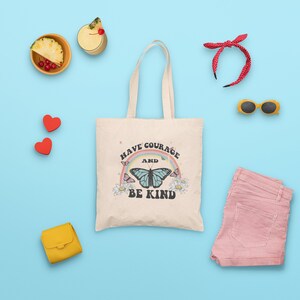May include: A beige tote bag with a rainbow and butterfly design. The bag has the text "Have Courage and Be Kind" printed on it.