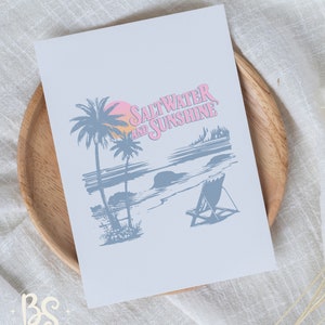 May include: A light blue print with a beach scene and the text "Saltwater and Sunshine" in pink. The scene includes palm trees, a beach, a sunset, and a beach chair.