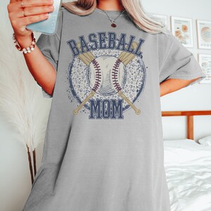 Op de afbeelding: Een grijs oversized T-shirt met een grafisch ontwerp van een honkbal, gekruiste knuppels en de tekst "Baseball Mom" in een blauw en wit lettertype. De grafiek is omgeven door een witte en blauwe luipaardprintcirkel.