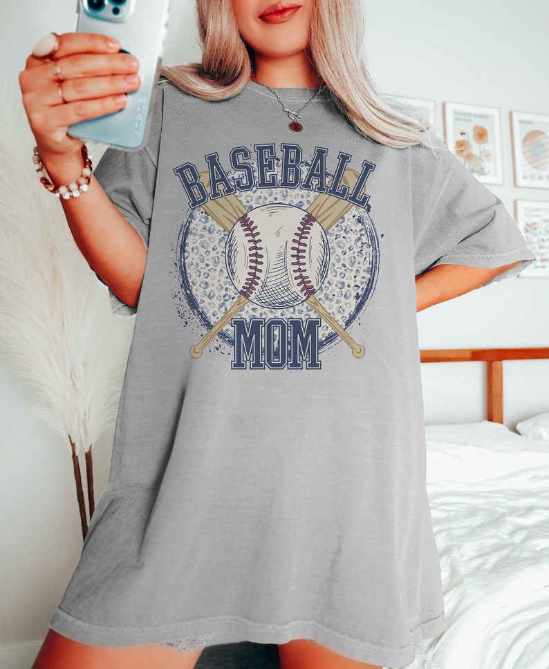 Op de afbeelding: Een grijs oversized T-shirt met een grafisch ontwerp van een honkbal, gekruiste knuppels en de tekst "Baseball Mom" in een blauw en wit lettertype. De grafiek is omgeven door een witte en blauwe luipaardprintcirkel.