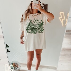 May include: A white oversized t-shirt with a green graphic of a skeleton sitting with a cup of coffee and surrounded by plants. The text on the shirt reads "Coffee & Plants".