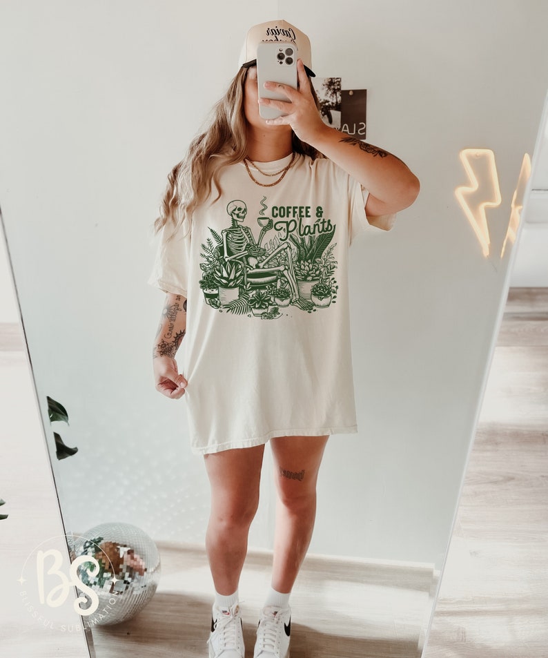May include: A white oversized t-shirt with a green graphic of a skeleton sitting with a cup of coffee and surrounded by plants. The text on the shirt reads "Coffee & Plants".