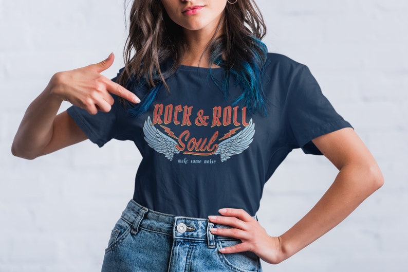 Puede incluir: Una camiseta azul marino con un gr&aacute;fico de estilo vintage que dice "Rock & Roll Soul" con alas y rayos. El texto debajo del gr&aacute;fico dice "make some noise".