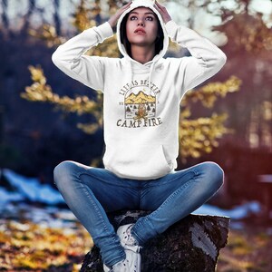 Pu&ograve; includere: Felpa con cappuccio bianca con una stampa grafica di una scena di fal&ograve; e il testo "Life is better by a campfire".