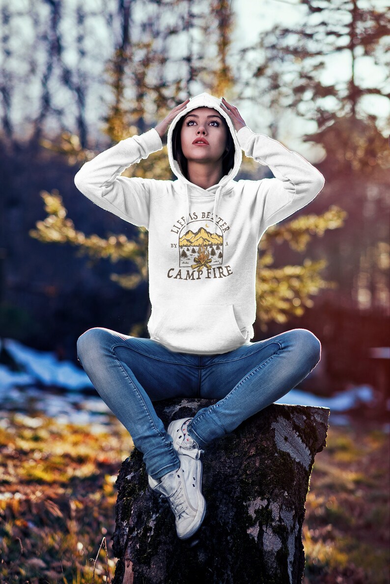 Pu&ograve; includere: Felpa con cappuccio bianca con una stampa grafica di una scena di fal&ograve; e il testo "Life is better by a campfire".