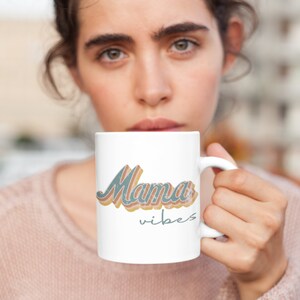 Puede incluir: Taza de cer&aacute;mica blanca con un dise&ntilde;o retro que presenta el texto "Mama vibes" en azul, naranja y rosa.