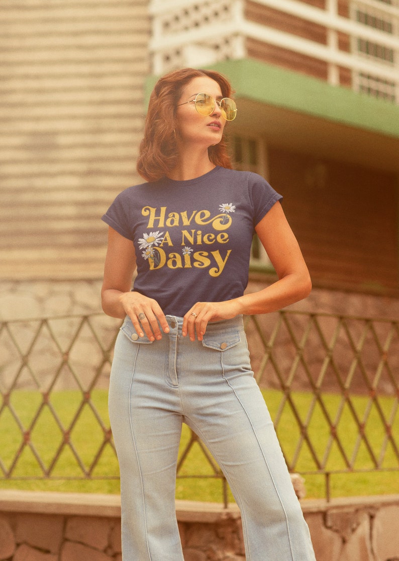 May include: A woman wearing a navy blue t-shirt with the text "Have A Nice Daisy" in yellow lettering and white daisies. She is wearing light blue jeans and yellow sunglasses.
