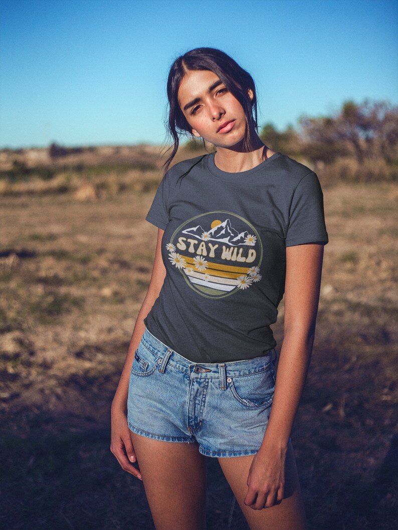 May include: A dark blue t-shirt with a graphic of mountains, a sun, and daisies. The graphic has the text "Stay Wild" in a yellow circle.