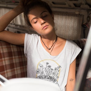 以下が含まれることがあります： 目を閉じた女性が、格子模様の座席のある車に座っています。彼女は、花柄のデザインと「WILD FLOWER」という文字が書かれた白いTシャツを着ています。彼女は2つのネックレスをしています。