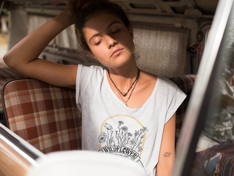 以下が含まれることがあります： 目を閉じた女性が、格子模様の座席のある車に座っています。彼女は、花柄のデザインと「WILD FLOWER」という文字が書かれた白いTシャツを着ています。彼女は2つのネックレスをしています。