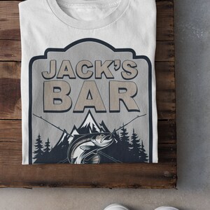 May include: White t-shirt with a graphic of a fishing sign that says "Jack's Bar" with a trout jumping out of the water and mountains in the background.