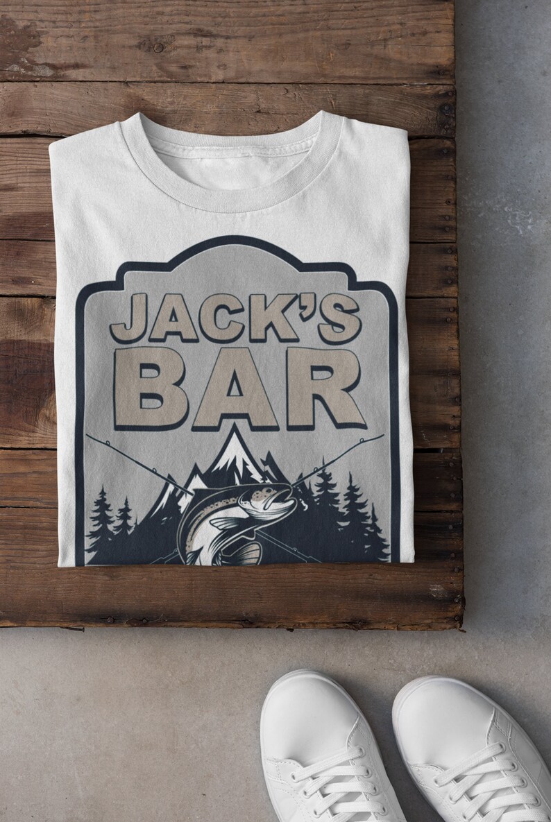May include: White t-shirt with a graphic of a fishing sign that says "Jack's Bar" with a trout jumping out of the water and mountains in the background.