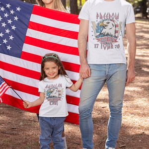 May include: A family of three wearing white t-shirts with American flag and eagle graphics. The shirts have the text "Freedom Born Free 17 Tour 76" and "American Born Free Kid". The family is standing in front of an American flag.