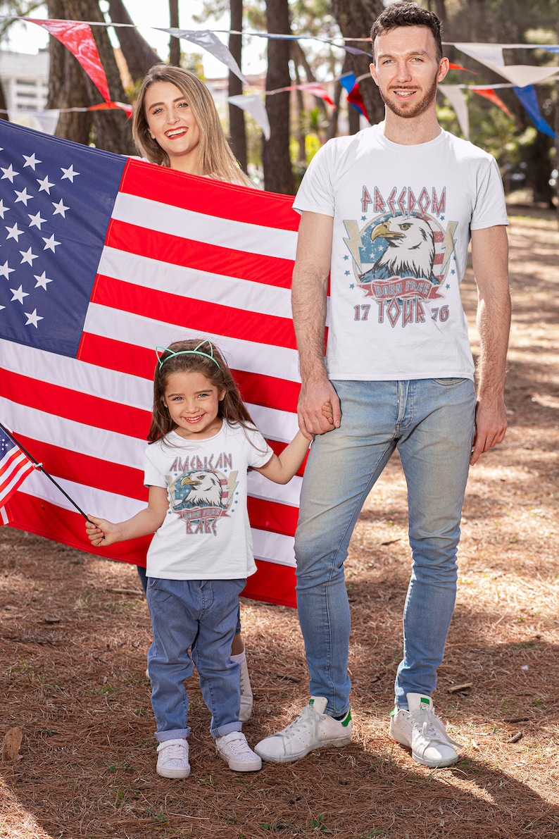 May include: A family of three wearing white t-shirts with American flag and eagle graphics. The shirts have the text "Freedom Born Free 17 Tour 76" and "American Born Free Kid". The family is standing in front of an American flag.