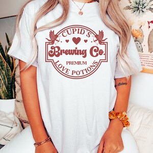 May include: White t-shirt with a red graphic of a circle with the text "Cupid's Brewing Co. Premium Love Potions" and two cherubs with arrows.