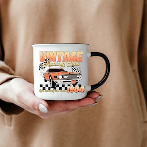 a hand holding a mug with the vintage racing club design. the retro design features a orange muscle car with checkered racing flags.  the words vintage racing club above and the words life in the fast lane 1984 below.