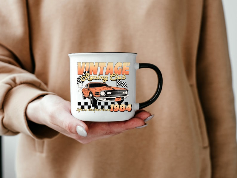 a hand holding a mug with the vintage racing club design. the retro design features a orange muscle car with checkered racing flags.  the words vintage racing club above and the words life in the fast lane 1984 below.