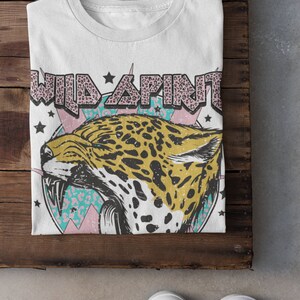 May include: White t-shirt featuring a graphic print of a jaguar head with a pink, blue and white background. The text "WILD SPIRIT" is printed in pink letters with a leopard print pattern.