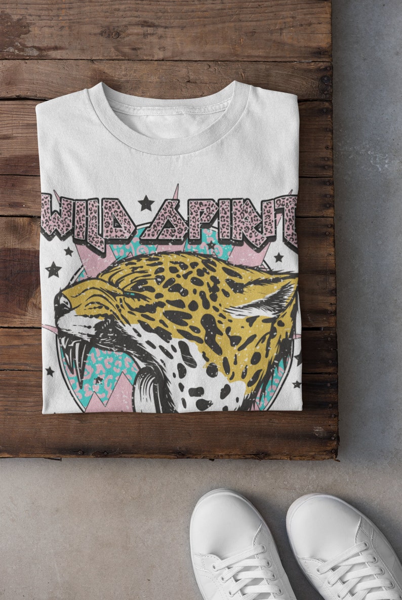 May include: White t-shirt featuring a graphic print of a jaguar head with a pink, blue and white background. The text "WILD SPIRIT" is printed in pink letters with a leopard print pattern.