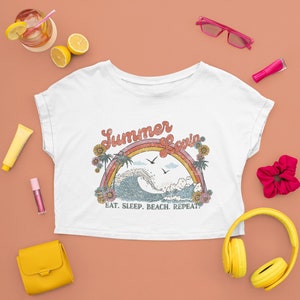 May include: White crop top with a retro design featuring a rainbow, ocean wave, palm trees, and the words "Summer Lovin'" and "Eat. Sleep. Beach. Repeat." The shirt is surrounded by accessories like sunglasses, headphones, and a purse.