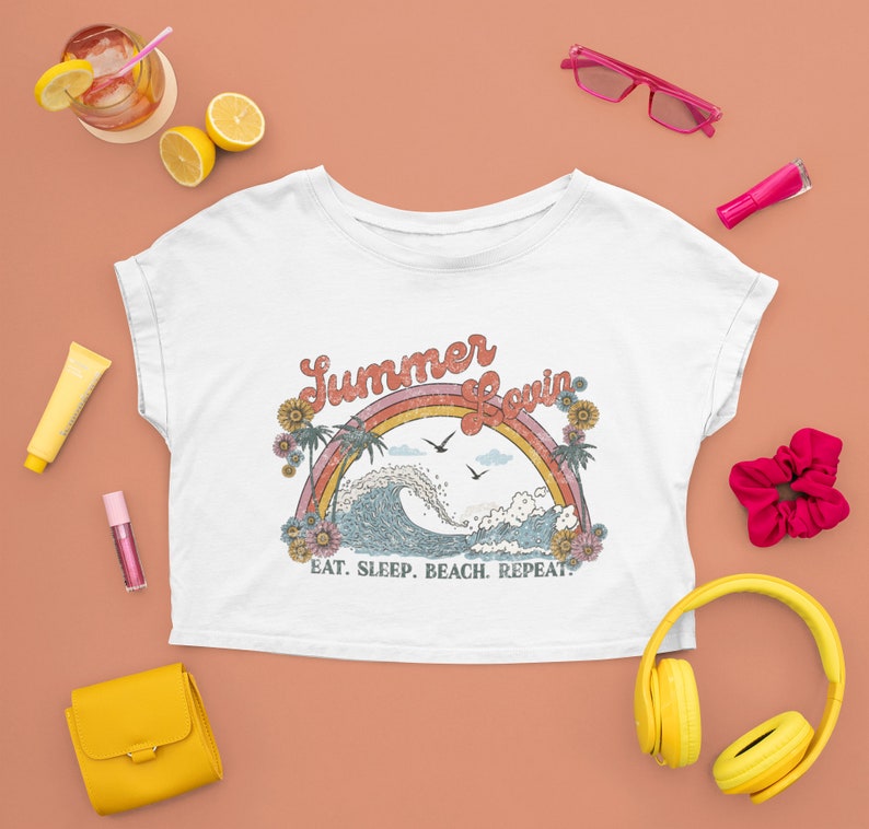 May include: White crop top with a retro design featuring a rainbow, ocean wave, palm trees, and the words "Summer Lovin'" and "Eat. Sleep. Beach. Repeat." The shirt is surrounded by accessories like sunglasses, headphones, and a purse.