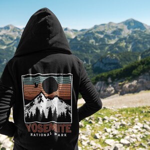 May include: Black hooded sweatshirt with a vintage-style graphic print of a mountain range, a sun, and a bird. The graphic is in shades of brown, orange, blue, and white. The text "Yosemite National Park" is printed below the graphic.