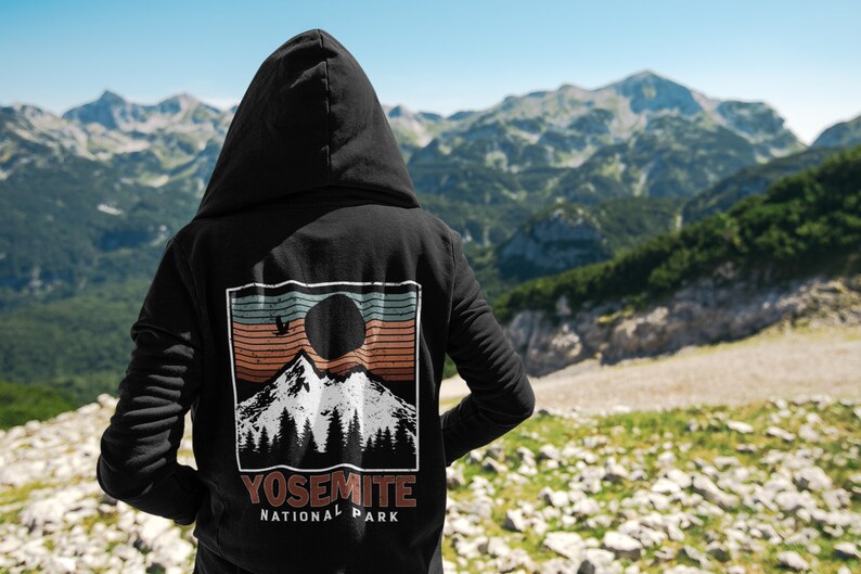 May include: Black hooded sweatshirt with a vintage-style graphic print of a mountain range, a sun, and a bird. The graphic is in shades of brown, orange, blue, and white. The text "Yosemite National Park" is printed below the graphic.