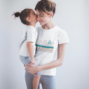 May include: A mother and daughter wearing matching white t-shirts. The daughter is being held by the mother. The mother's shirt has the word "MAMA" printed on it with a heart and colourful stripes.