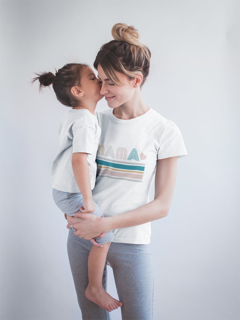 May include: A mother and daughter wearing matching white t-shirts. The daughter is being held by the mother. The mother's shirt has the word "MAMA" printed on it with a heart and colourful stripes.