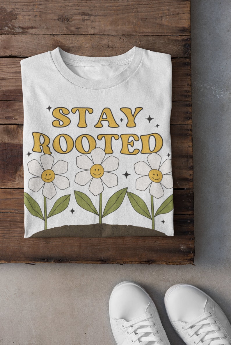 May include: White t-shirt with the text "STAY ROOTED" in yellow letters. Three white daisies with yellow centers and smiley faces are below the text.