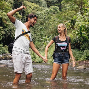 May include: A couple laughing and holding hands while wading in a shallow stream. The man is wearing a white t-shirt with a colorful graphic and the woman is wearing a black t-shirt with the text "River Life" in pink and blue.
