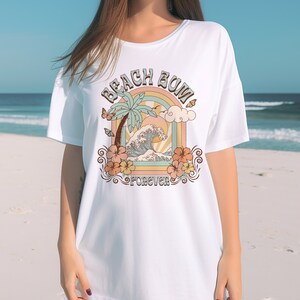 Pu&ograve; includere: Maglietta bianca oversize con una stampa grafica di una palma, onde oceaniche e un arcobaleno. Il testo "Beach Bum Forever" &egrave; stampato sulla maglietta.