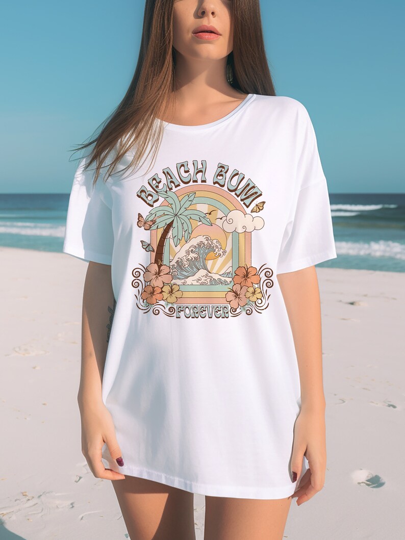 Pu&ograve; includere: Maglietta bianca oversize con una stampa grafica di una palma, onde oceaniche e un arcobaleno. Il testo "Beach Bum Forever" &egrave; stampato sulla maglietta.