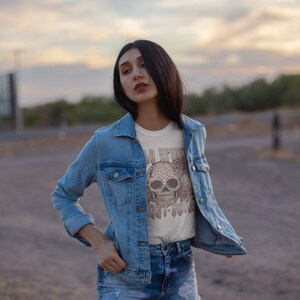 May include: A woman wearing a light blue denim jacket over a white t-shirt with a brown skull graphic and the text "Old Friends Get It Done".